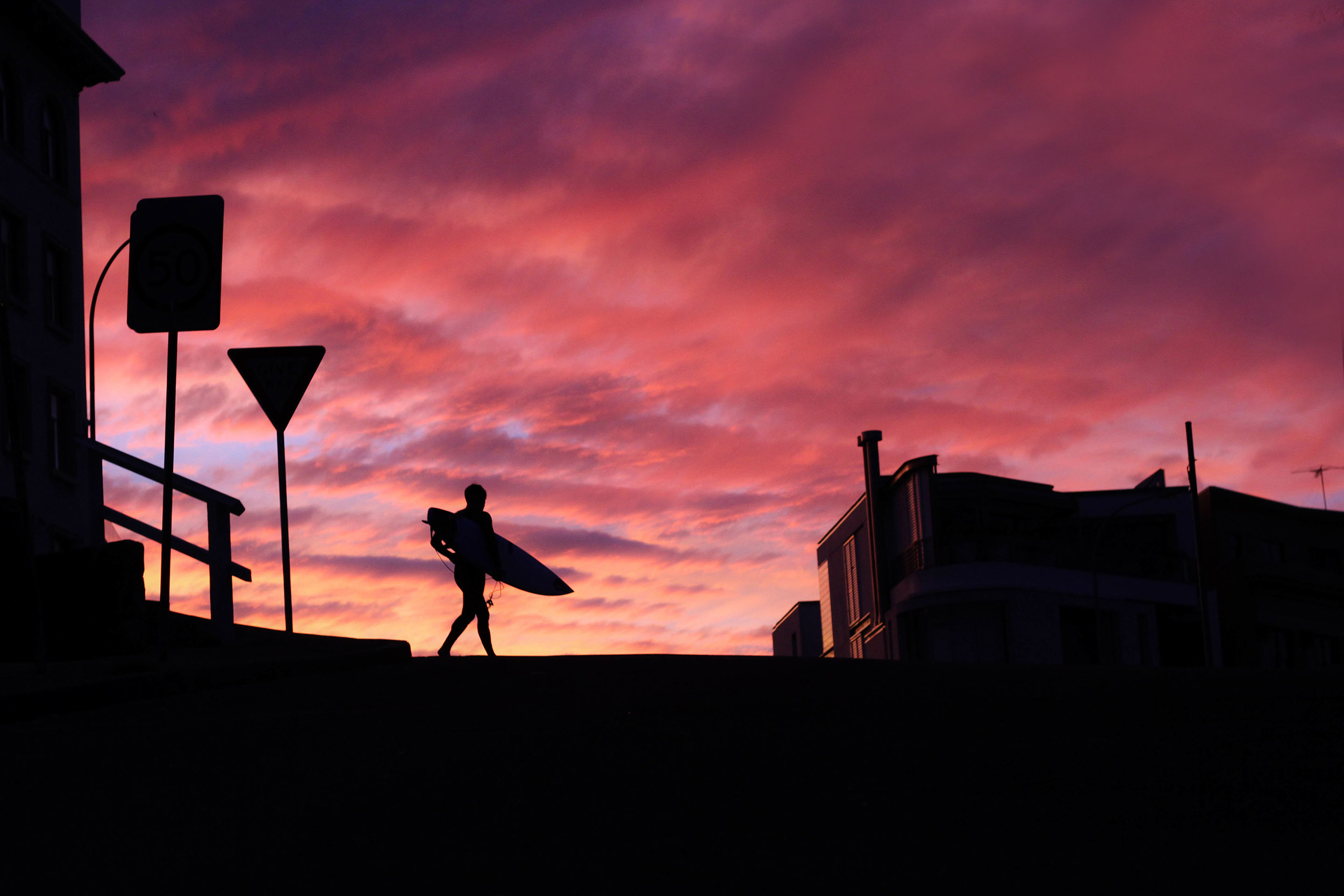 Surfer holding surfboard silhouetted against fiery red sunset sky walking across the pier in Bondi Beach