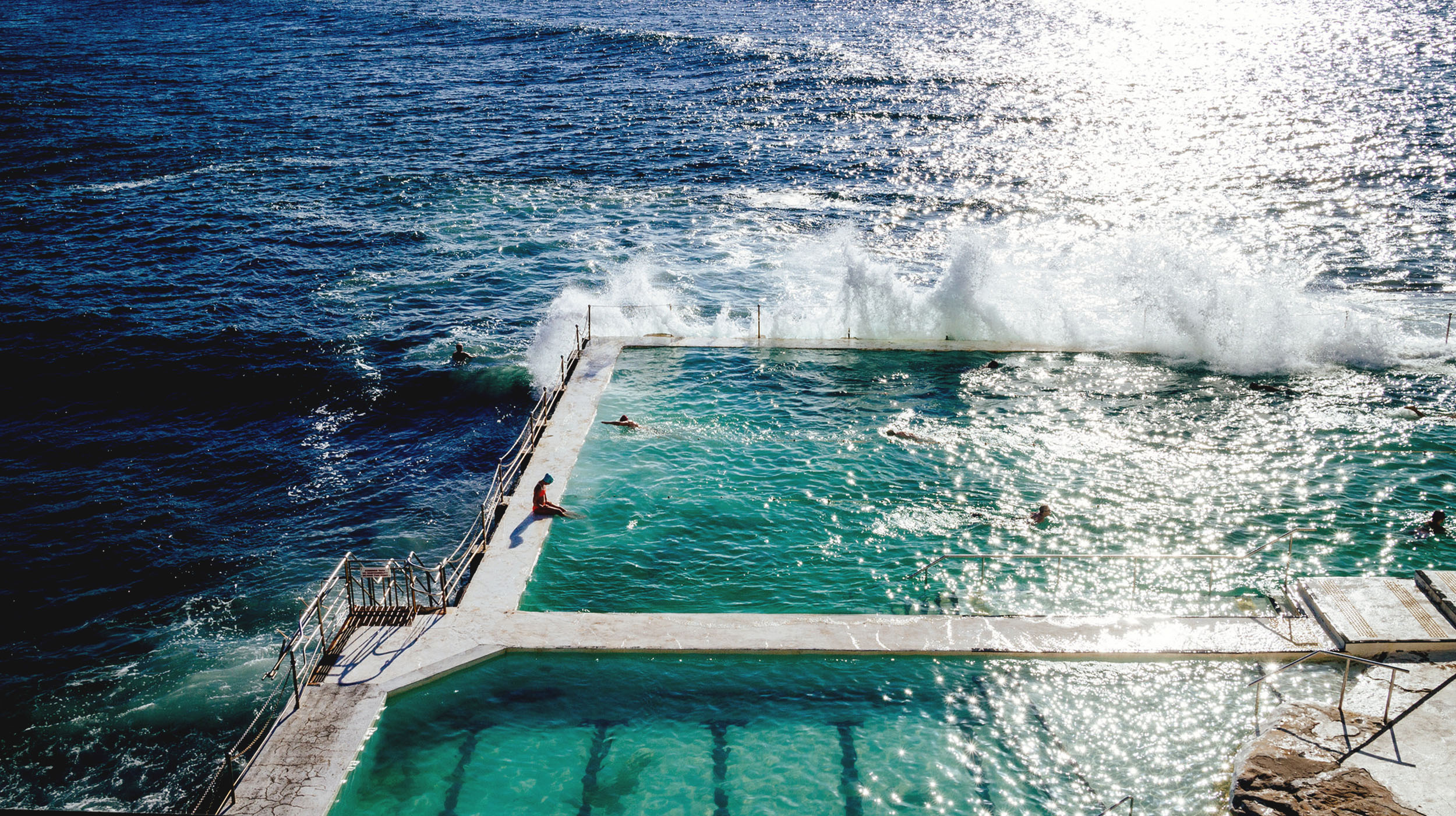 Sunny oceanside pools with waves crashing into the side and a person diving in 