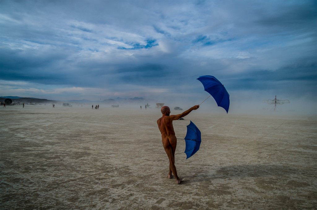 Nude man backing camera, walking through Black Rock desert waving two open blue umbrellas at the Burning Man festival