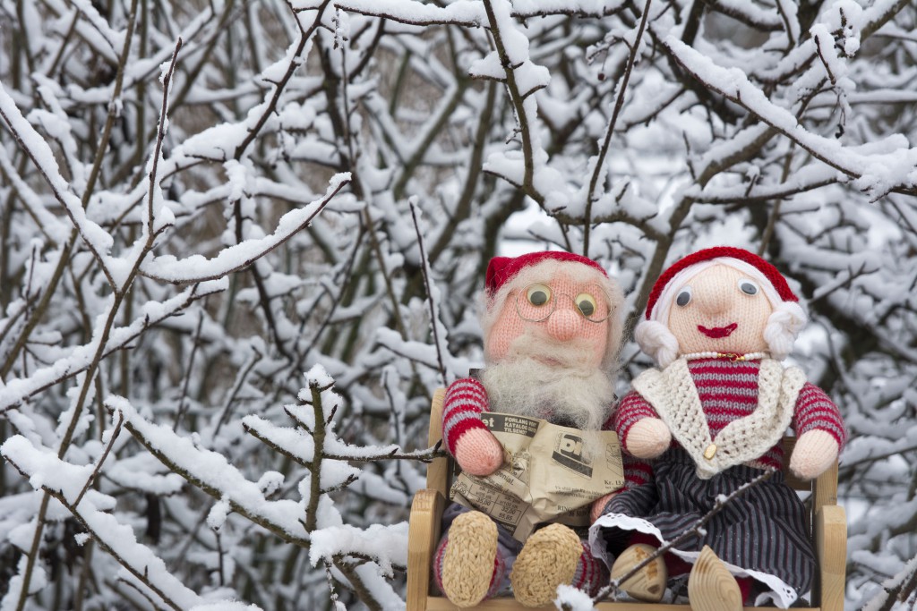 A pair of stuffed Santa’s Nisser dolls sitting on a bench amongst snowy twigs