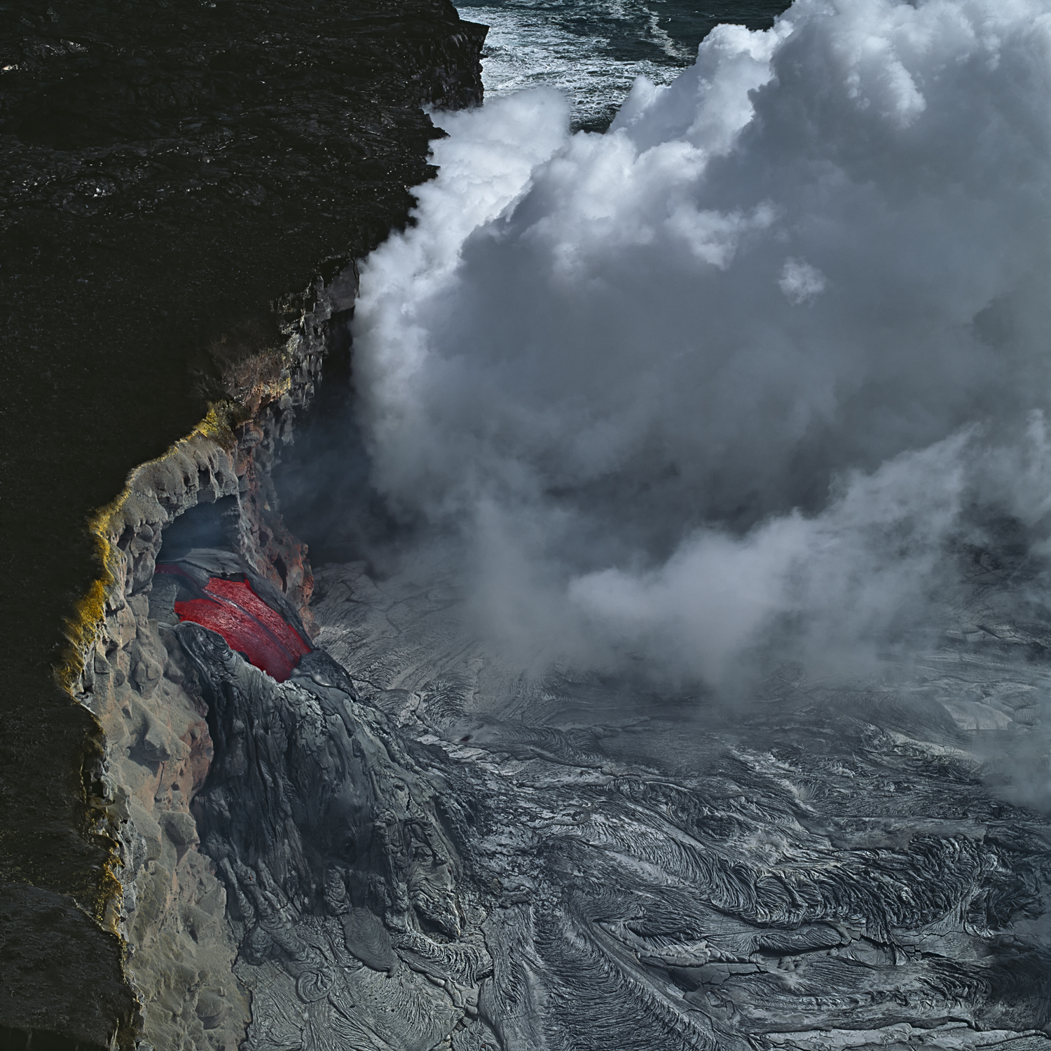 Red lava flowing down a craggy rockface from tunnels with steam clouds swirling around, Kilauea, Hawai’i 
