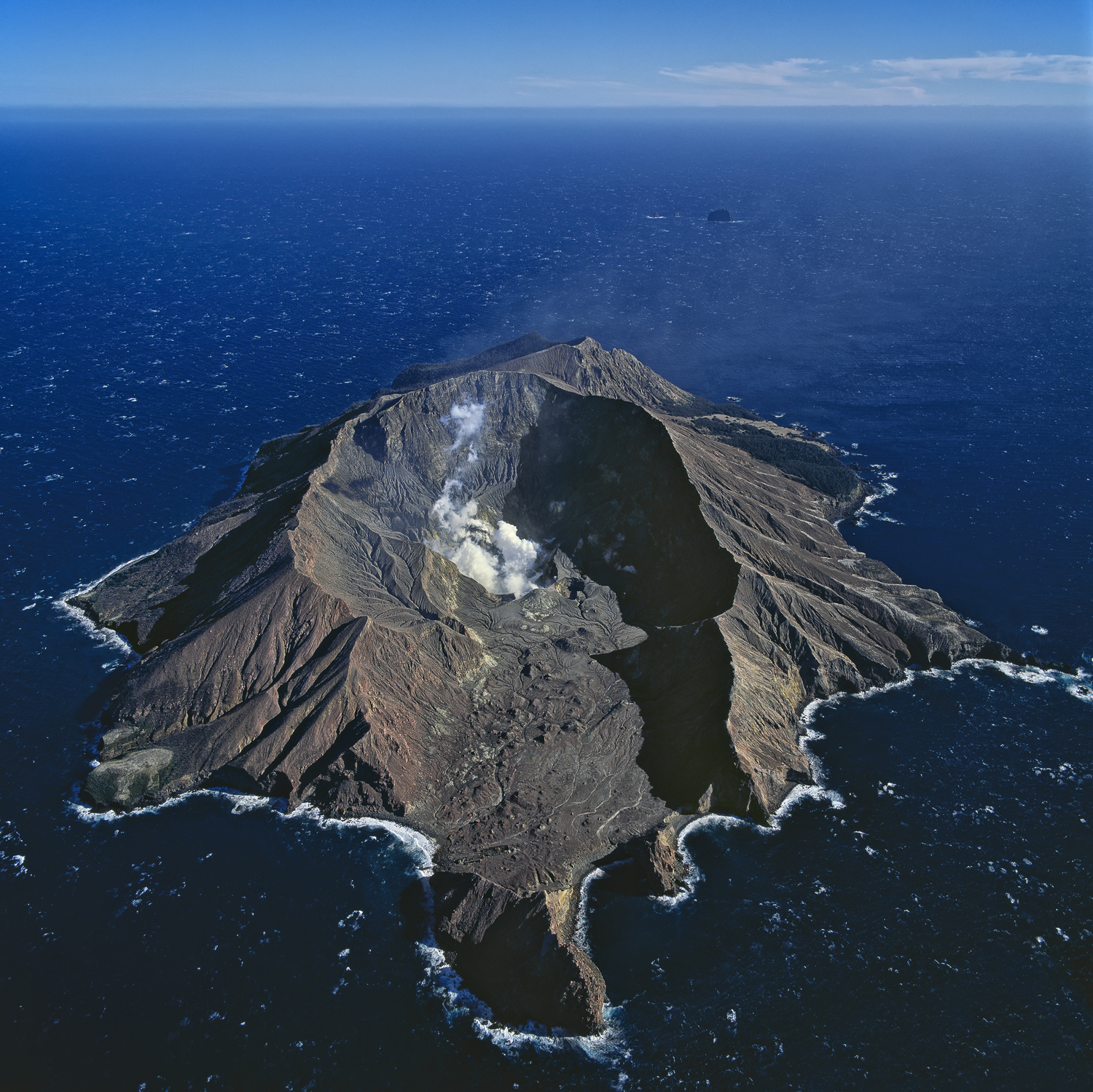 The rocky and striated horseshoe shaped tip of the White Island volcano projecting out of a deep blue sea, New Zealand.