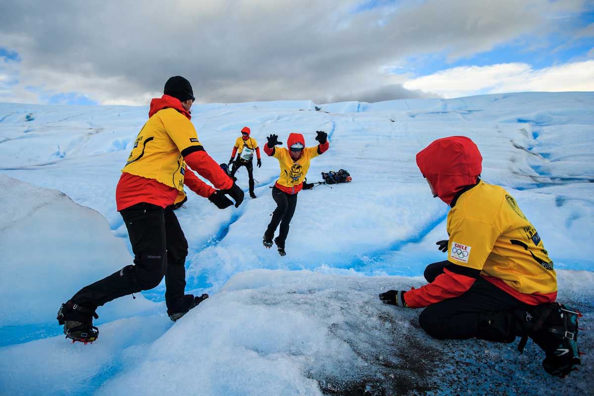 Group clad in yellow and red suits in icy snow and one of them jumping across a crevasse