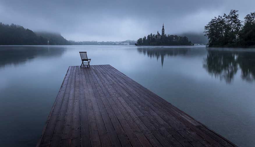 A chair at the end of a long wooden jettyin Lake Bled at dusk with a church on an island in the background at dusk