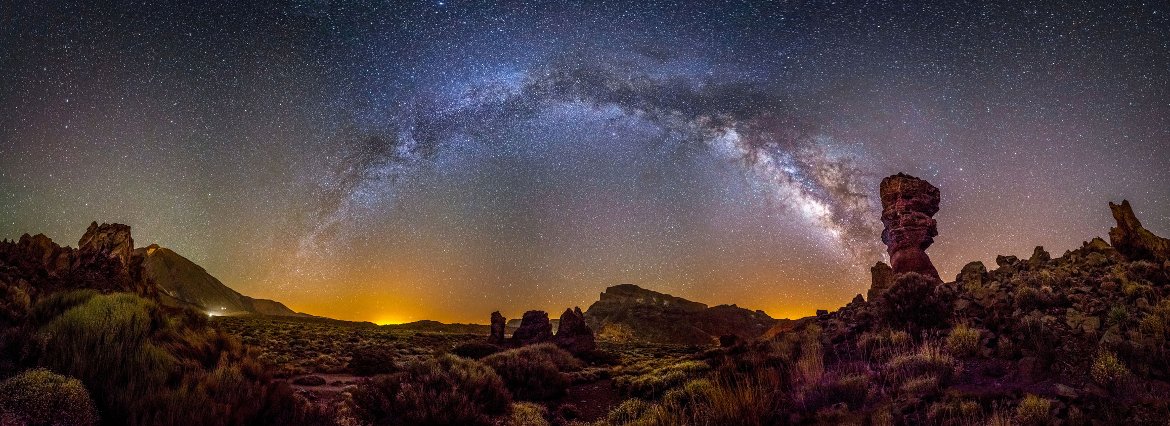 The Milky Way in a parabola over a very craggy and rocky landscape interspersed with tussock grass at night in Canary Islands, Spain