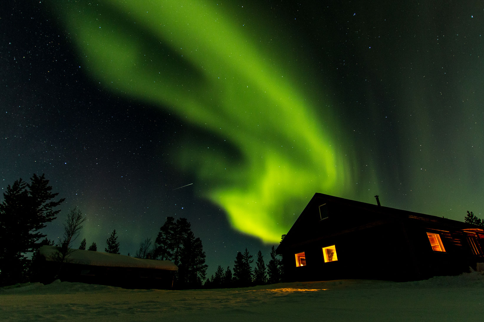 A house in snow at night, lighted up from within and a stunning bright green aurora formation in the sky behind it, Sweden