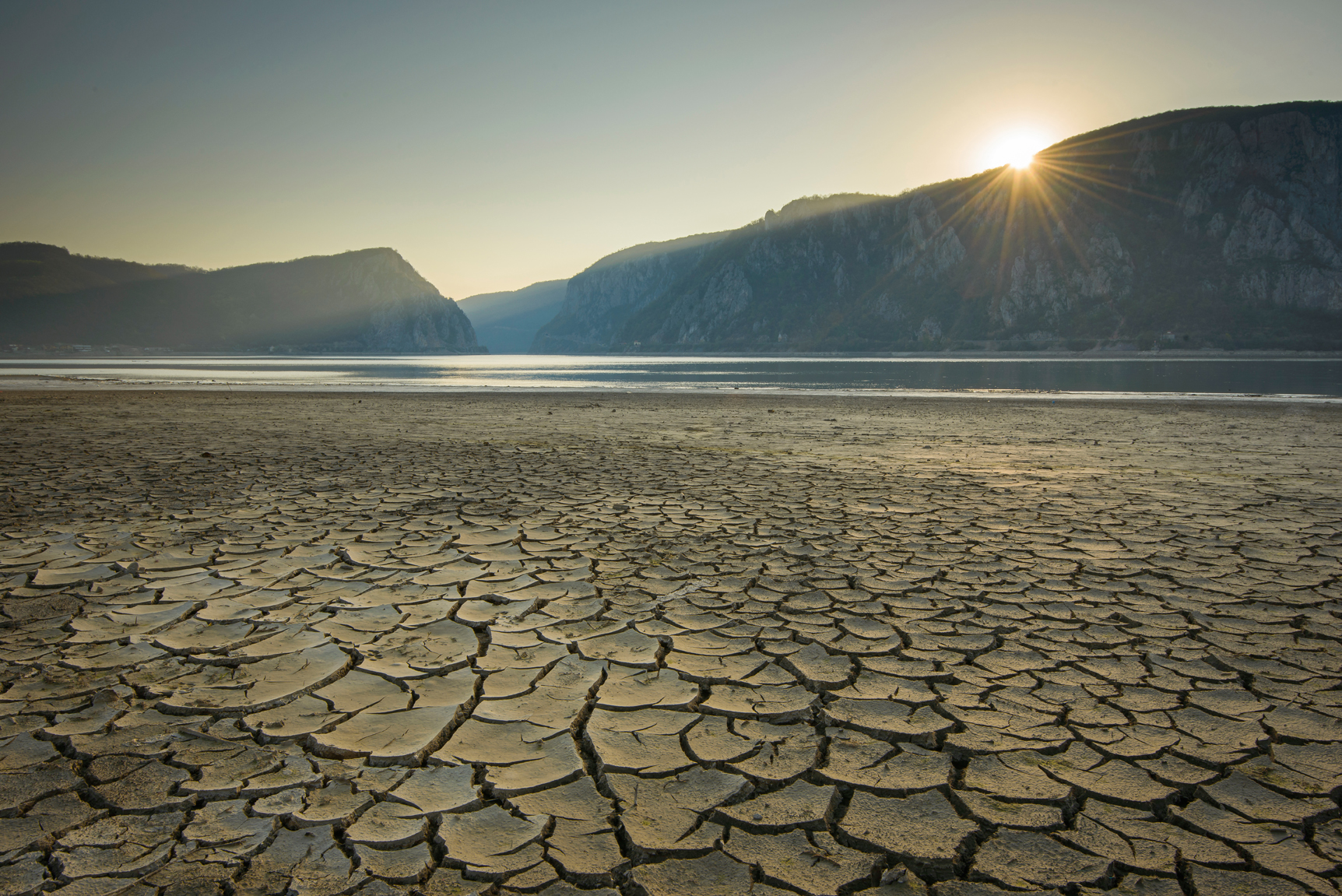 Wild World: Cracked and dry riverbed extending into the distance ending at the Danube River and mountains in the background and the sun just showing above them in Eastern Europe.