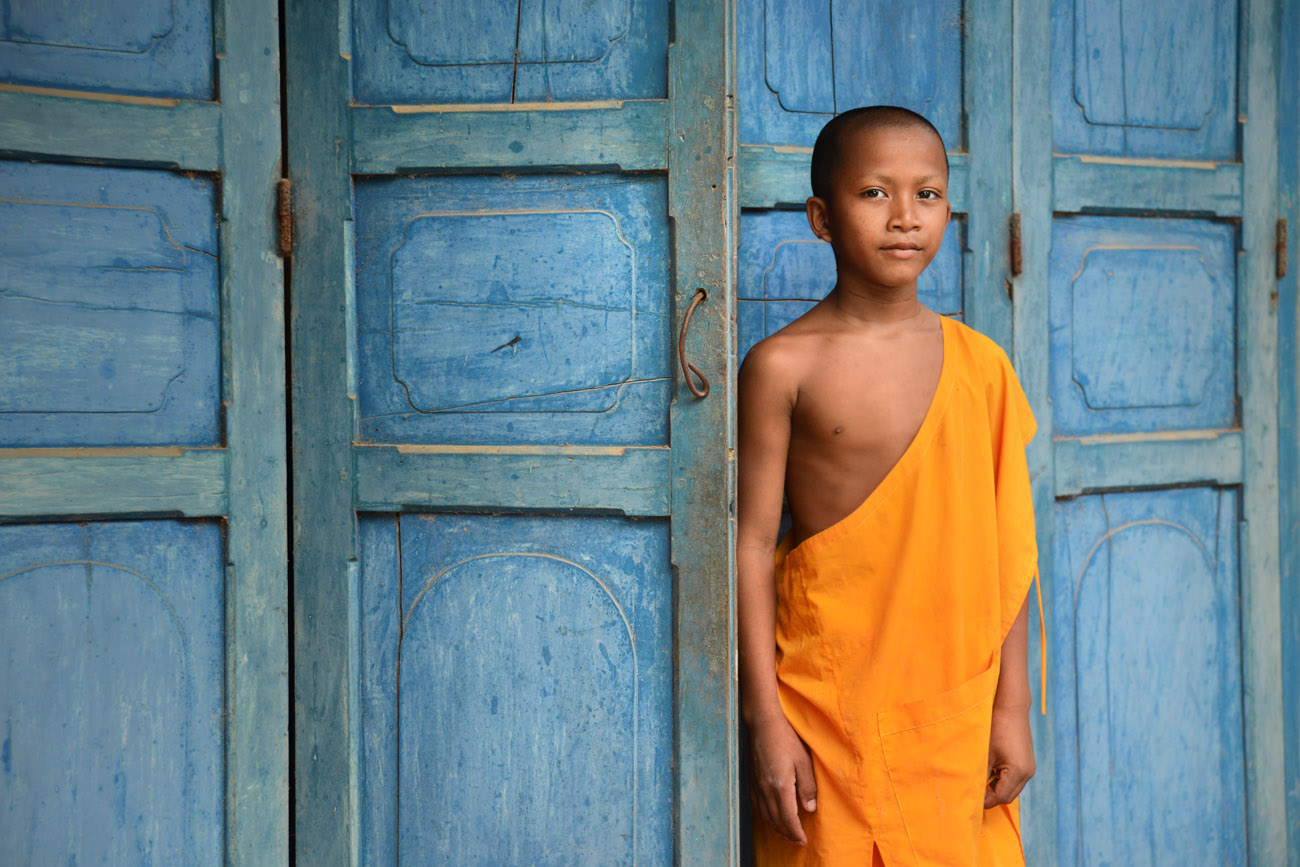 A novice monk in orange robes stands by the blue doors of a Khmer monastery in the south of Vietnam, near the Cambodian border