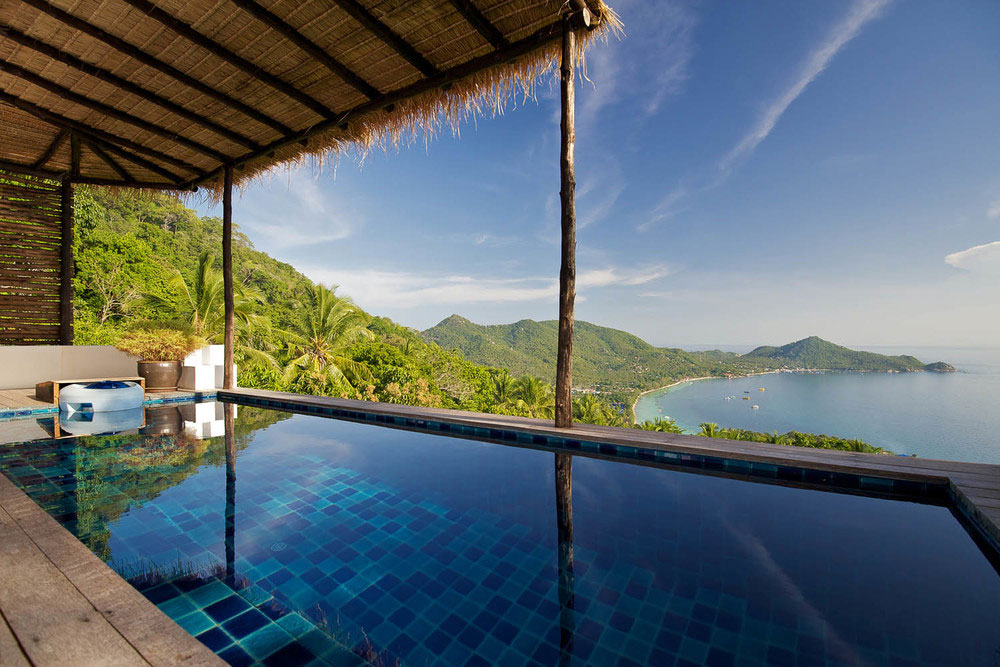 A view out from a Casas Del Sol's pool having a thatched roof down towards the lush vegetation, beach, sea and hills in the distance, Thailand