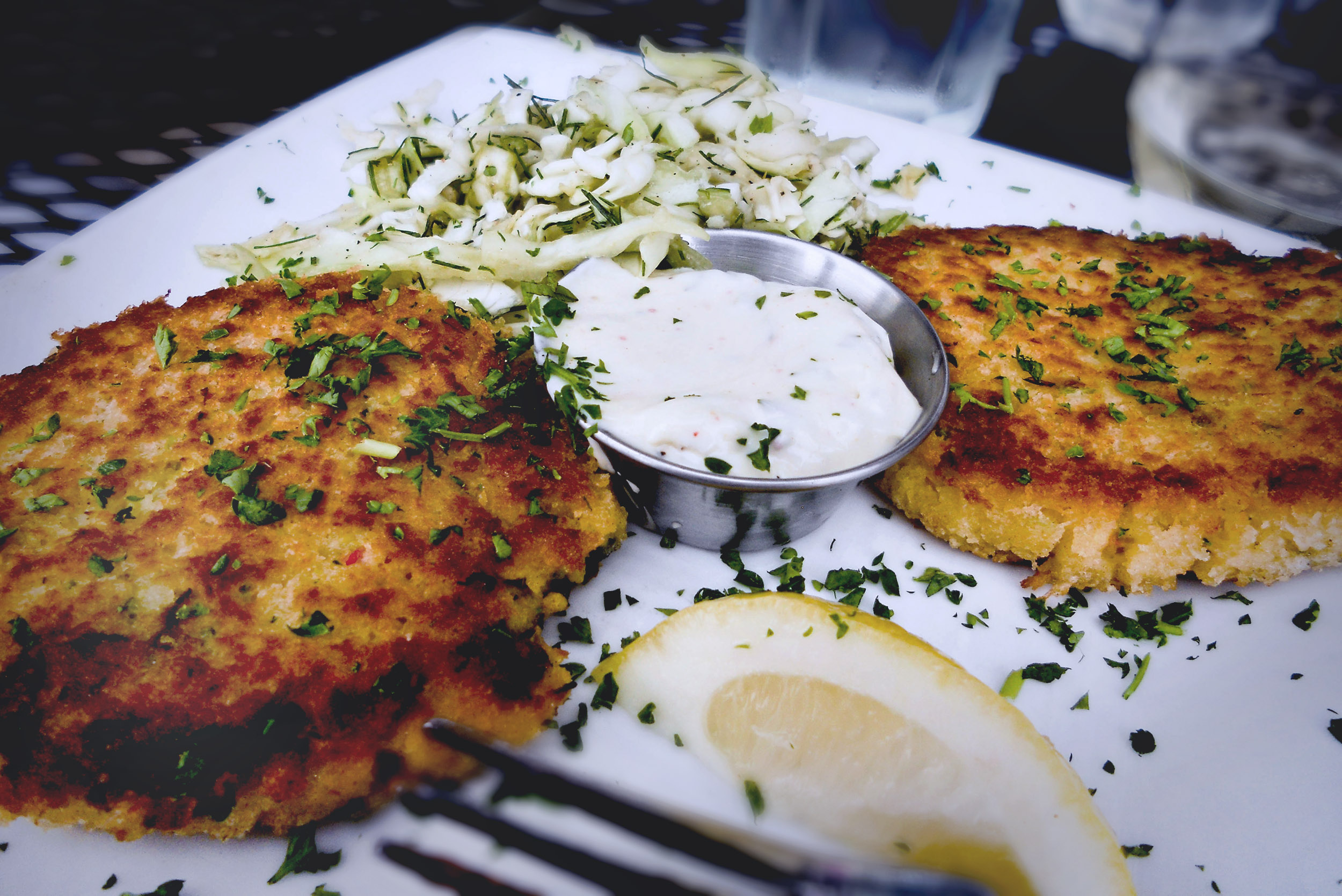 A platter with two dungeness crab cakes, fennel slaw, tartar sauce and a slice of lemon with a scattering of chopped chives.