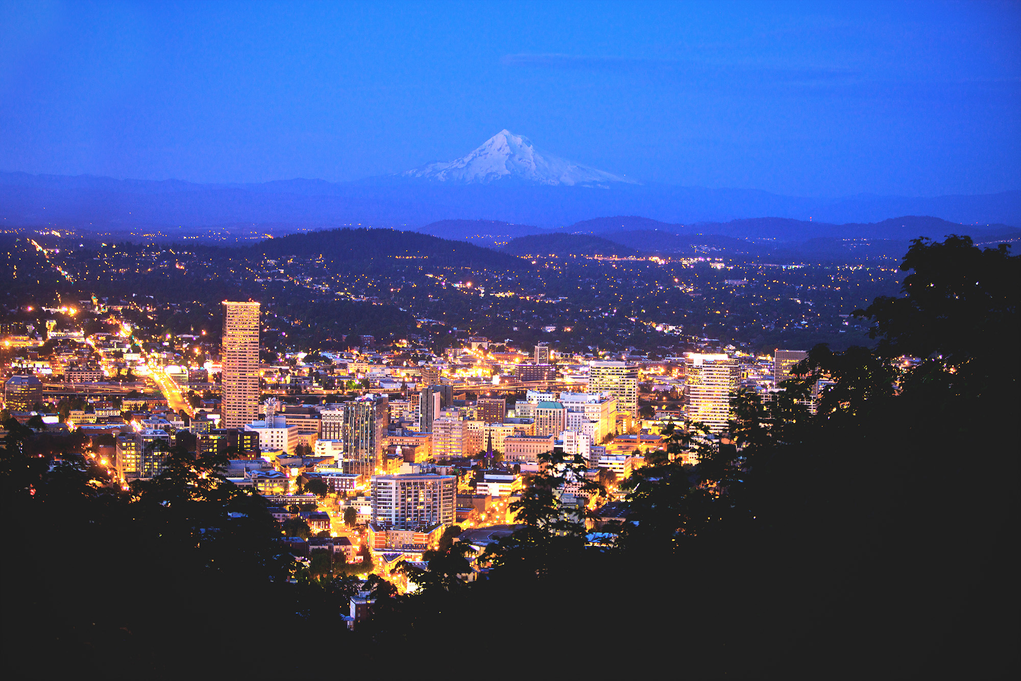 A lighted up Portland city with a snow capped Mt Hood dimly visible in the distant night sky