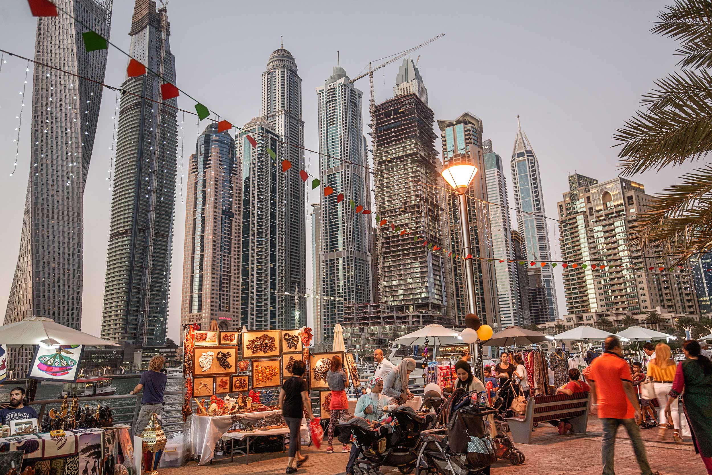 The lighted up stalls and vendors in the evening with people walking past them and the skyscrapers of Dubai in the background