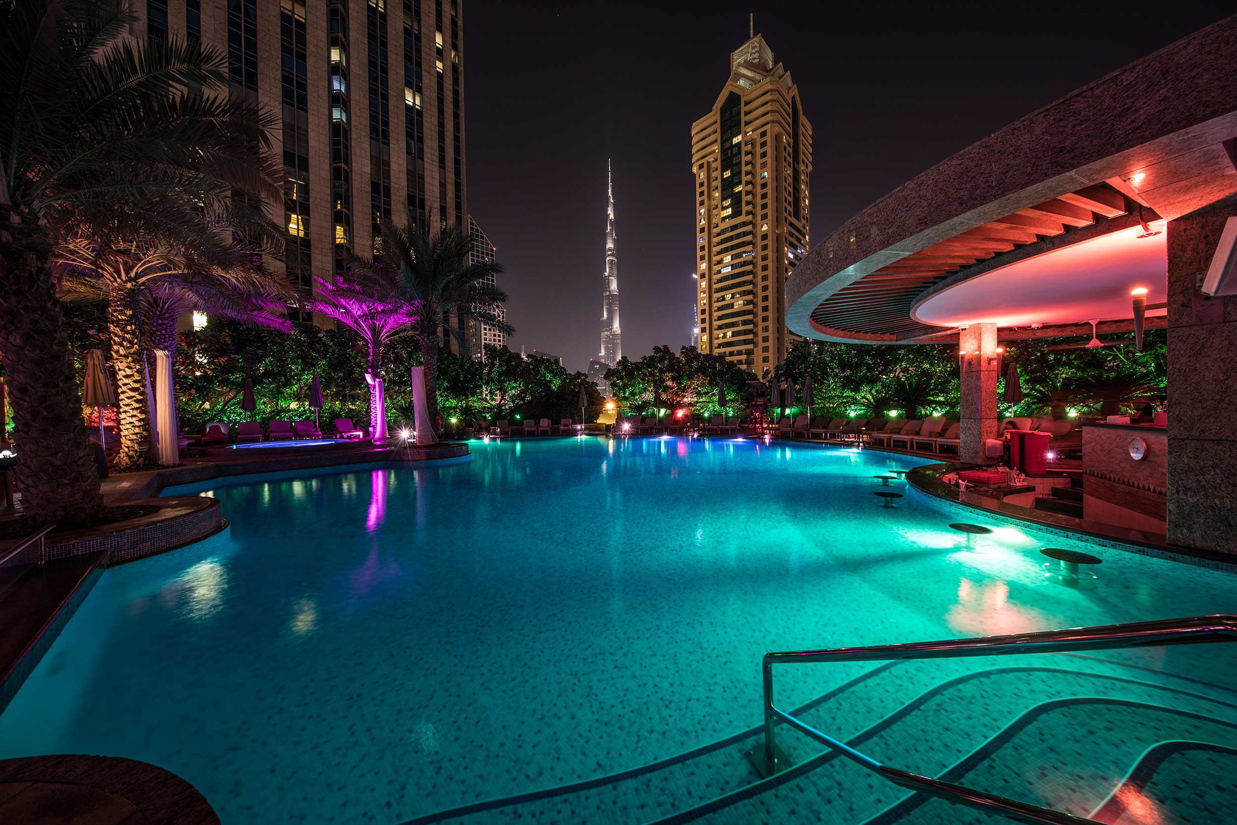 Multi-coloured lights streaming across a swimming pool at night with the lighted skyscrapers of dubai in the background