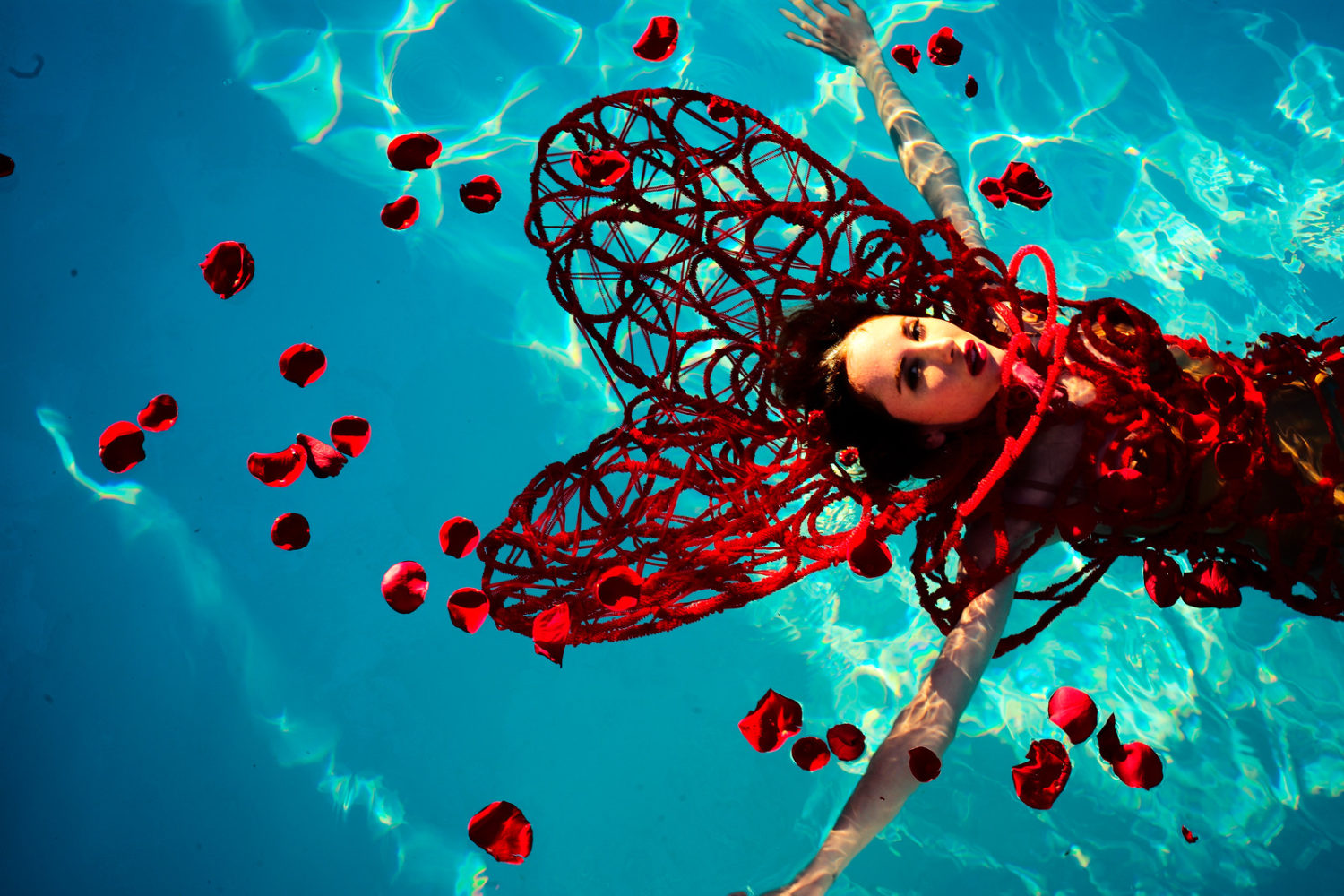 Girl in red wings and costume floating in a pool with arms stretched wide and rose petals scattered and floating around her