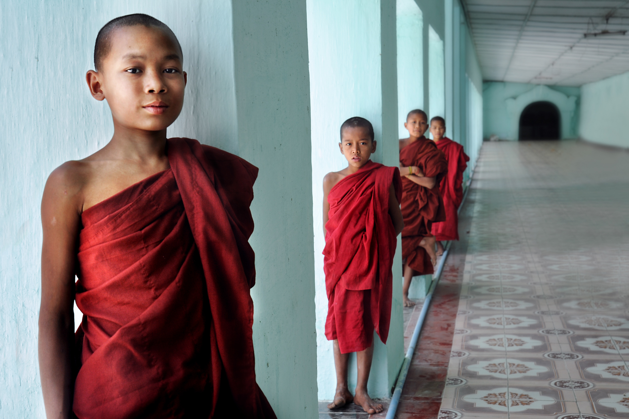 Four boy monks standing in a line each by a pillar down a corridor