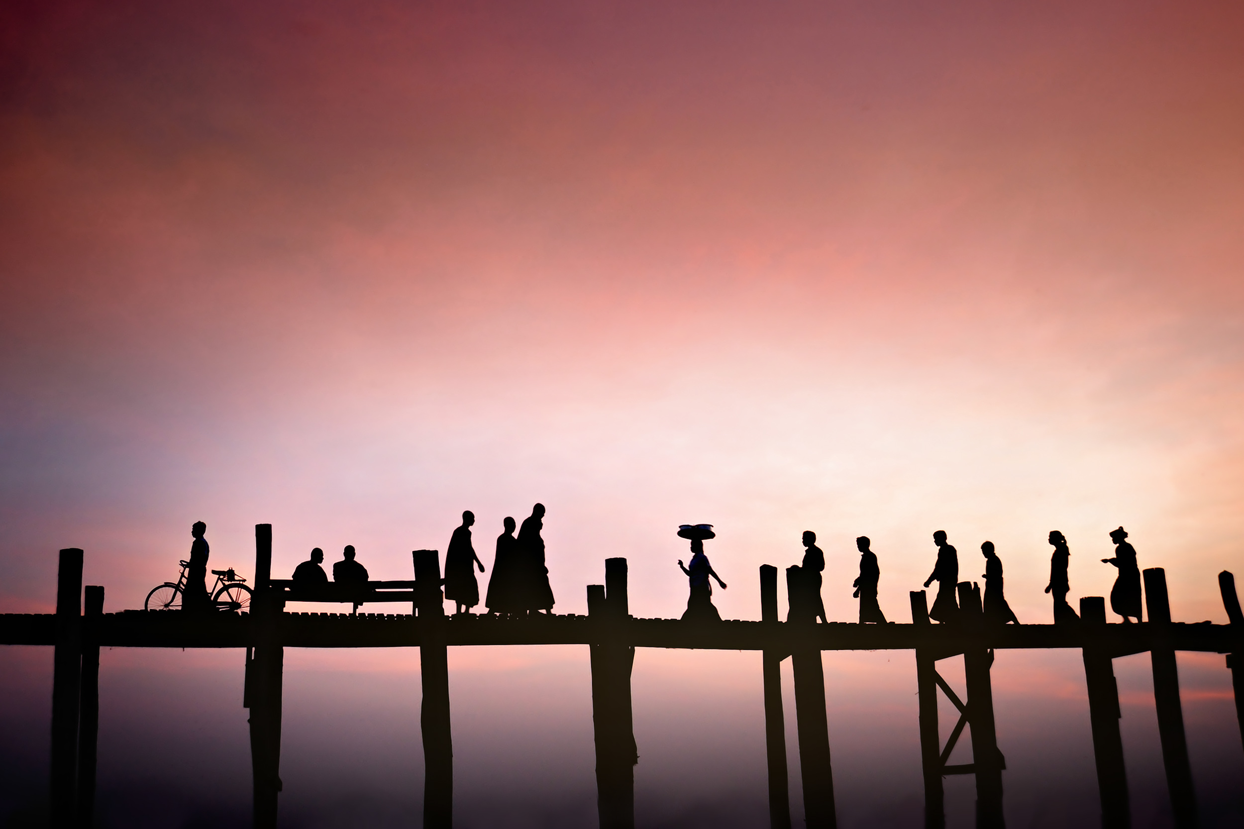 Silhouettes of multiple people crossing a wooden bridge on tree trunk poles at sunset
