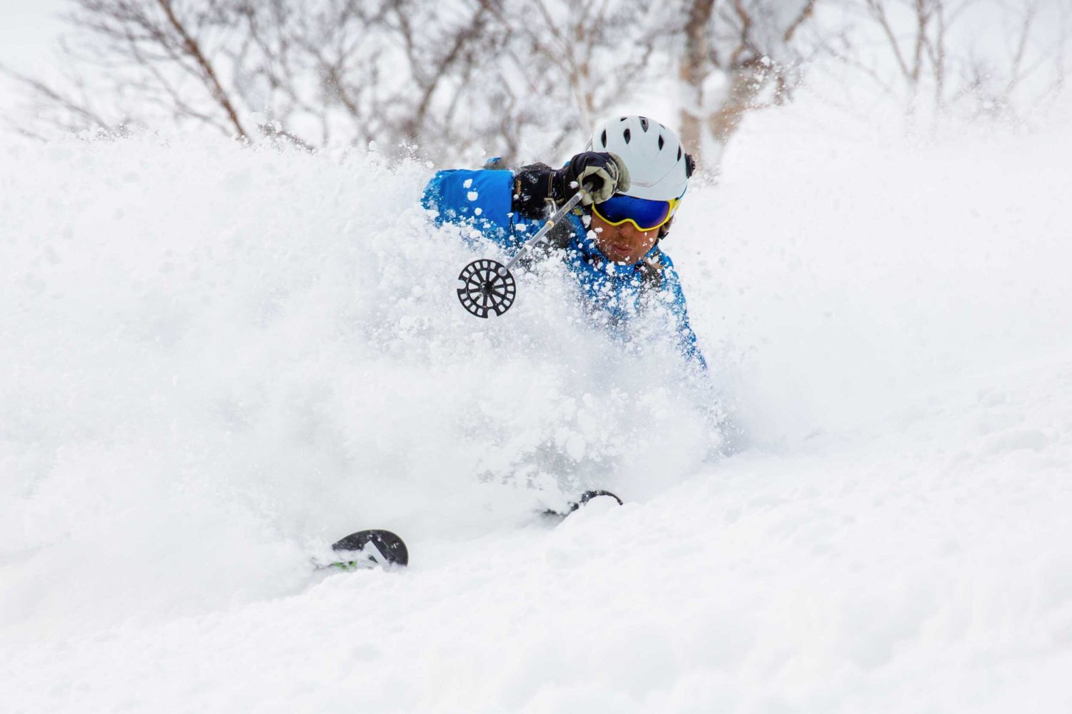A skier in blue and a white helmet throwing up a spray of snow on a turn in Niseko, Japan
