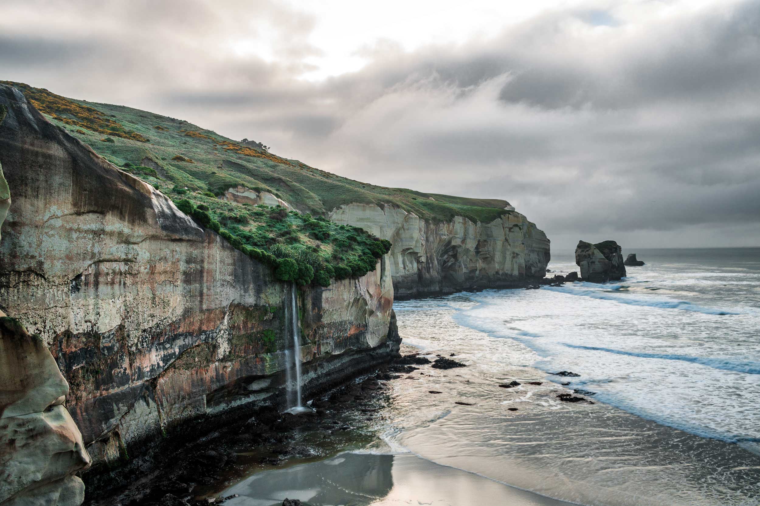 A cliff coastline with a grassy top and a stream of water falling off it onto the sand below, New Zealand
