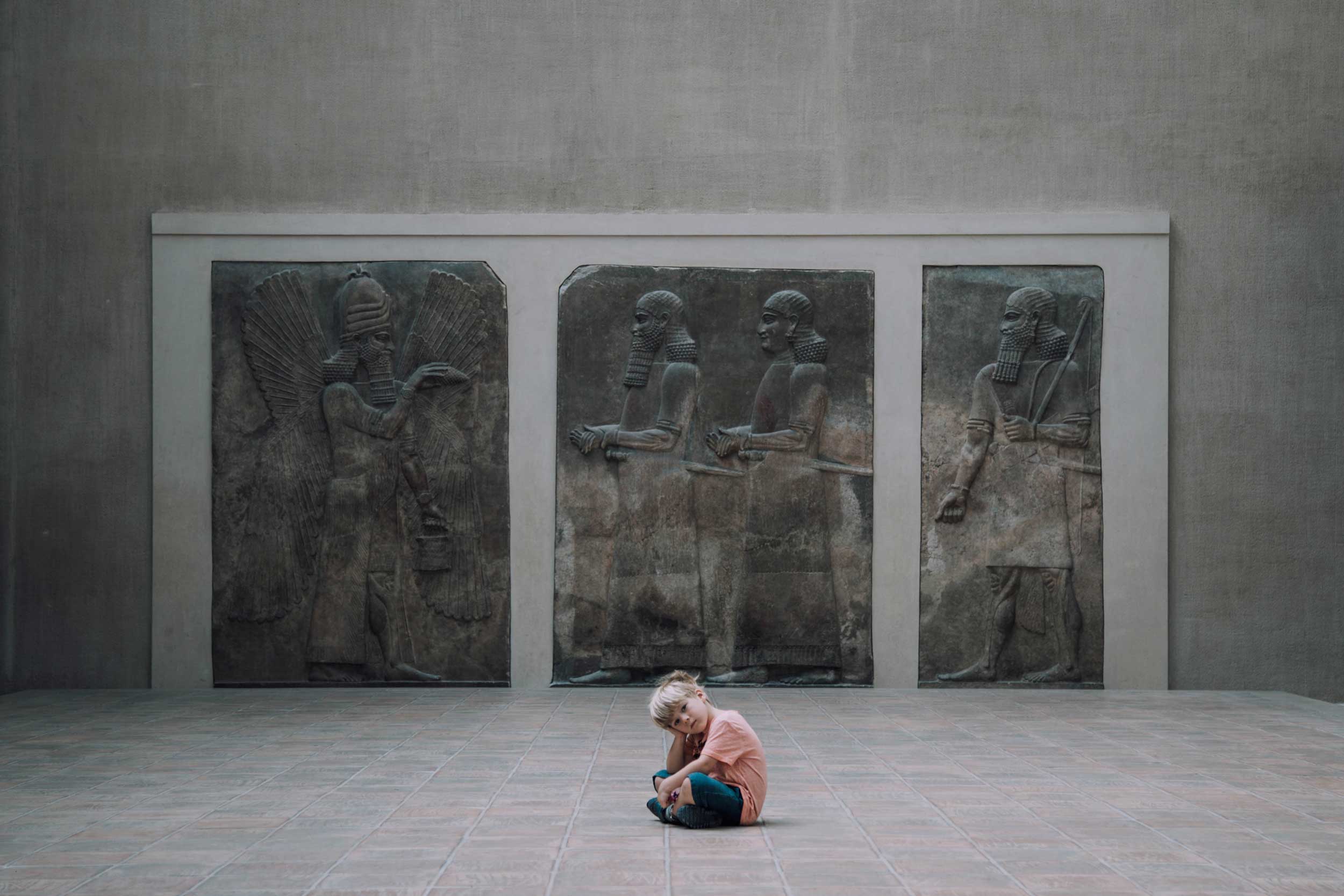 A small boy seated cross-legged staring into camera in front of a carved stone wall, Paris