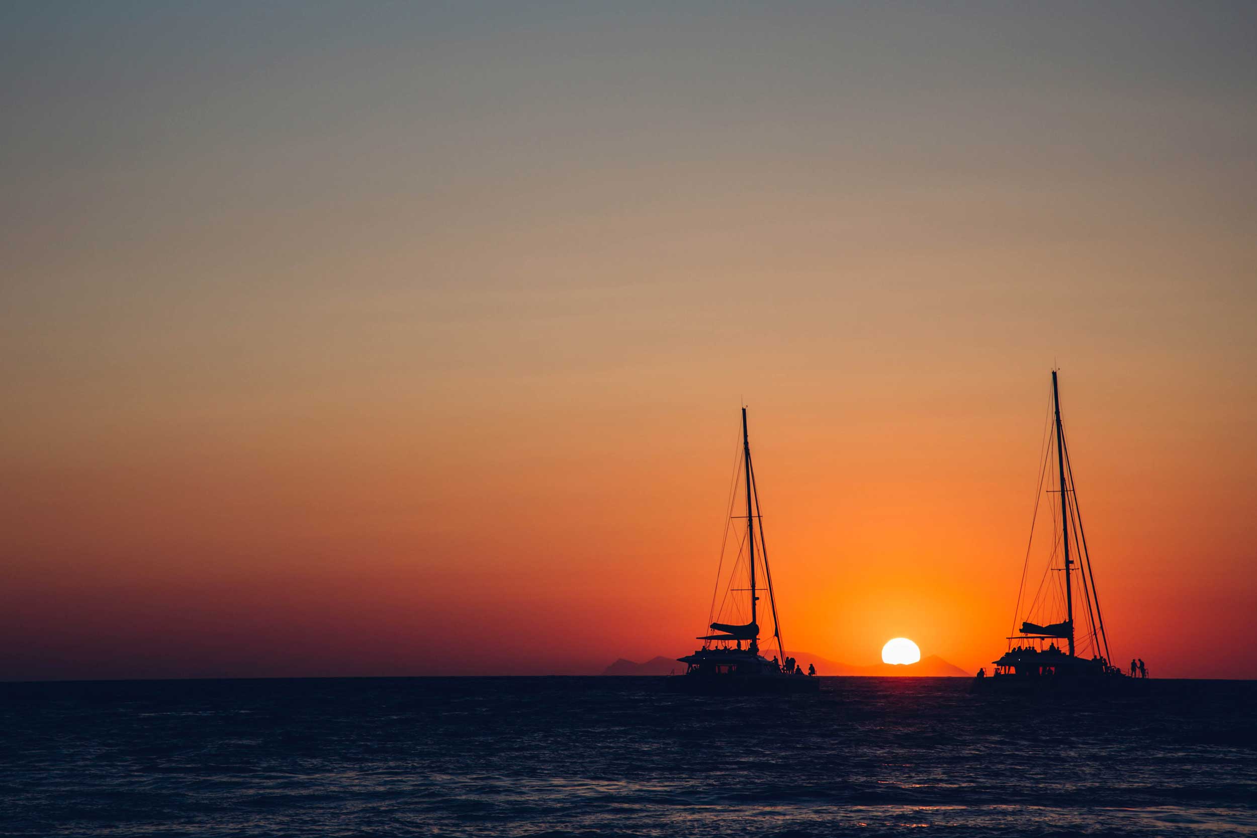 Two sailboats, sails down, silhouetted against a dark sea and orange tinted sky in Santorini, Greece