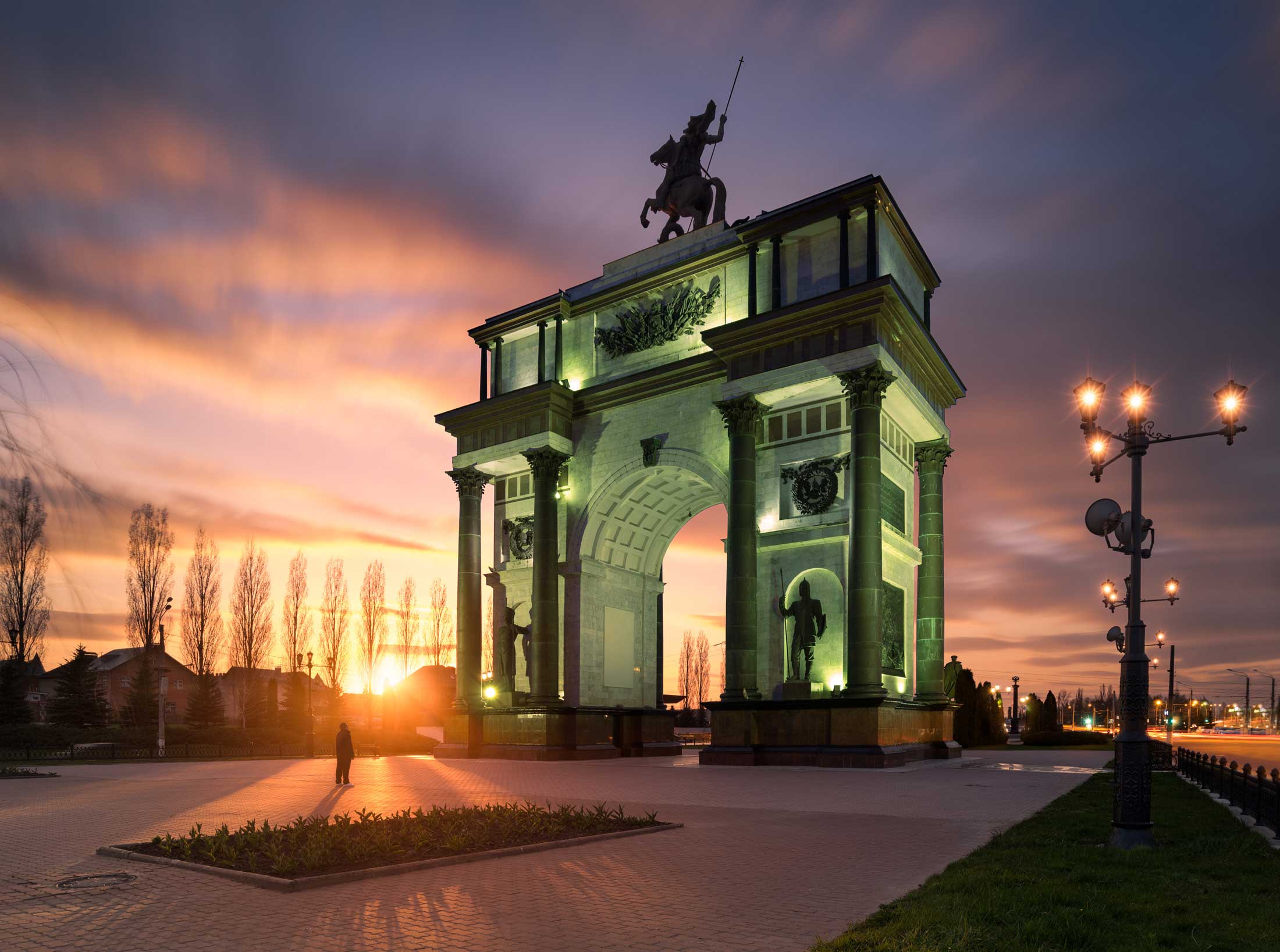 Man in a beam of light staring up at an elaborate arch with a horse and rider statue atop it, Kursk, Russia